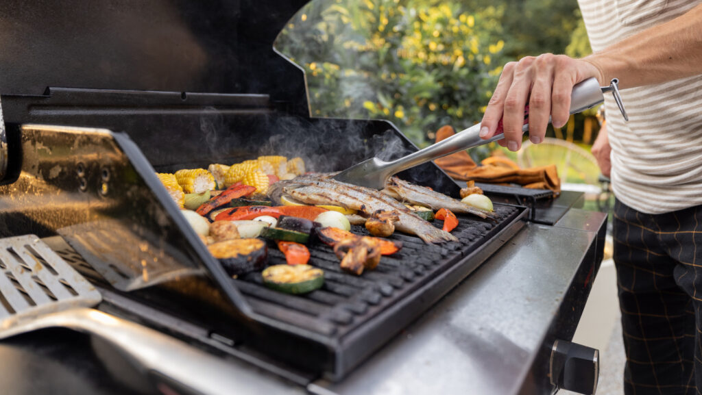 Man flips vegetables and meat on grill with tongs.